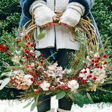 A woman showcasing festive winter wreath ideas, standing in the garden on a patch of snow, holding a foliage and berry wreath.