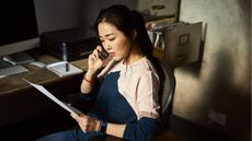A thoughtful-looking woman looks at paperwork while talking on the phone.