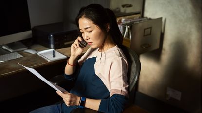 A thoughtful-looking woman looks at paperwork while talking on the phone.