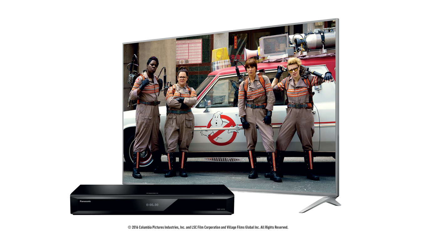 Panasonic DMP-UB700 4K Blu-ray player on a white background next to a TV displaying Ghostbusters