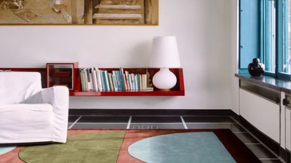 modern room with white walls, large artwork, blue window frames, floating red bookshelf with table lamp, white slipcover sofa, and colorful graphic rugs on a black glossy floors