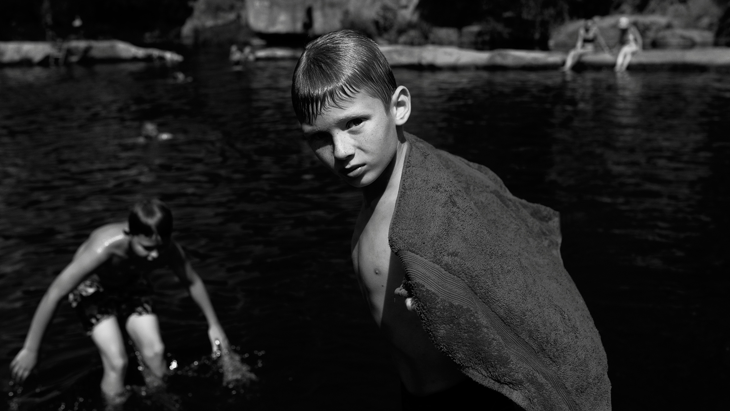 High contrast, black-and-white image, showing two boys at a lake, one is looking straight into the camera