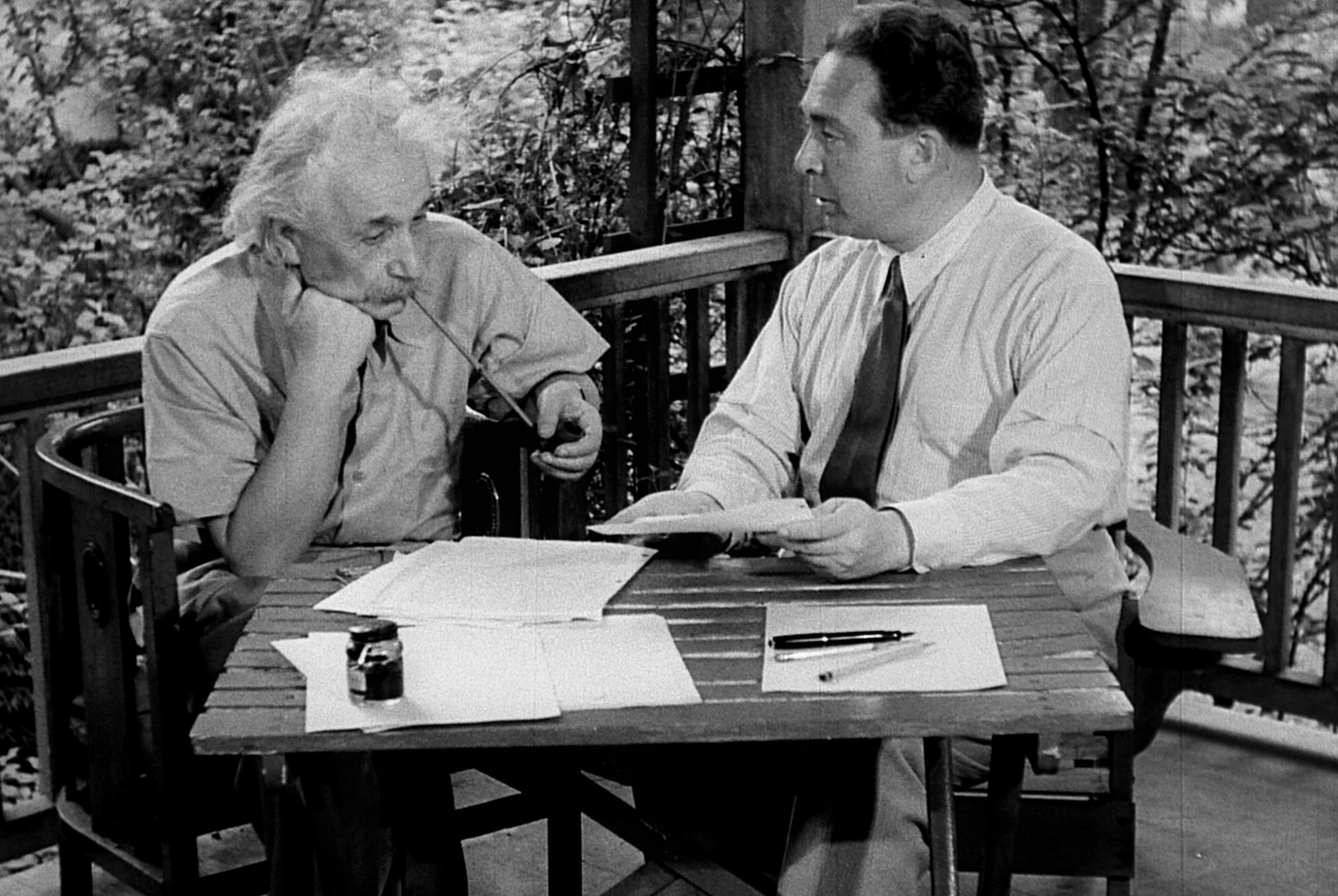 a man (albert einstein) smokes a long pipe while sitting at a desk on an outdoor porch, next to another man in a dress shirt and tie