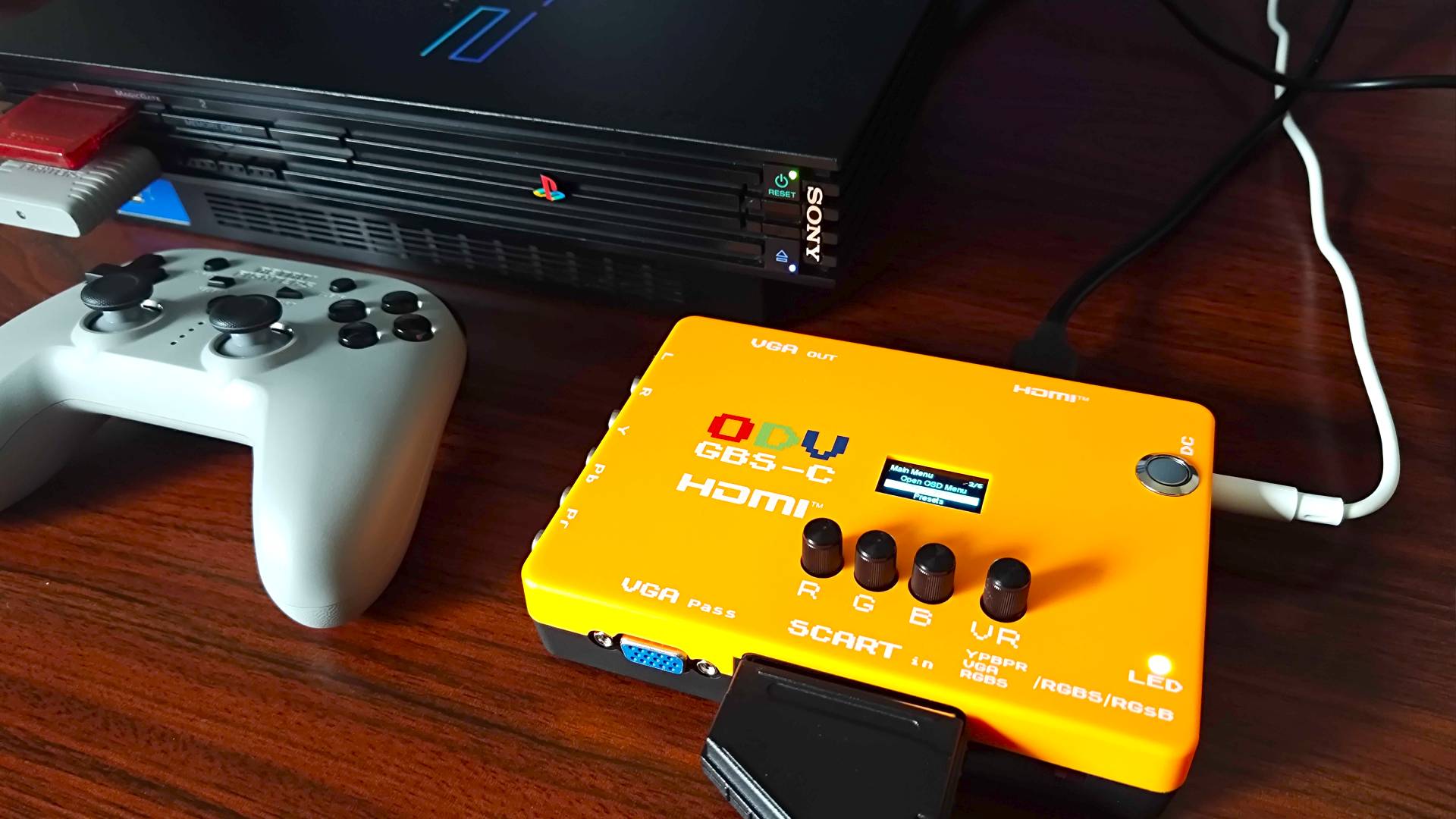 GBS-C upscaler next to PS2 and Retro Fighters Defender controller on woodgrain desk.