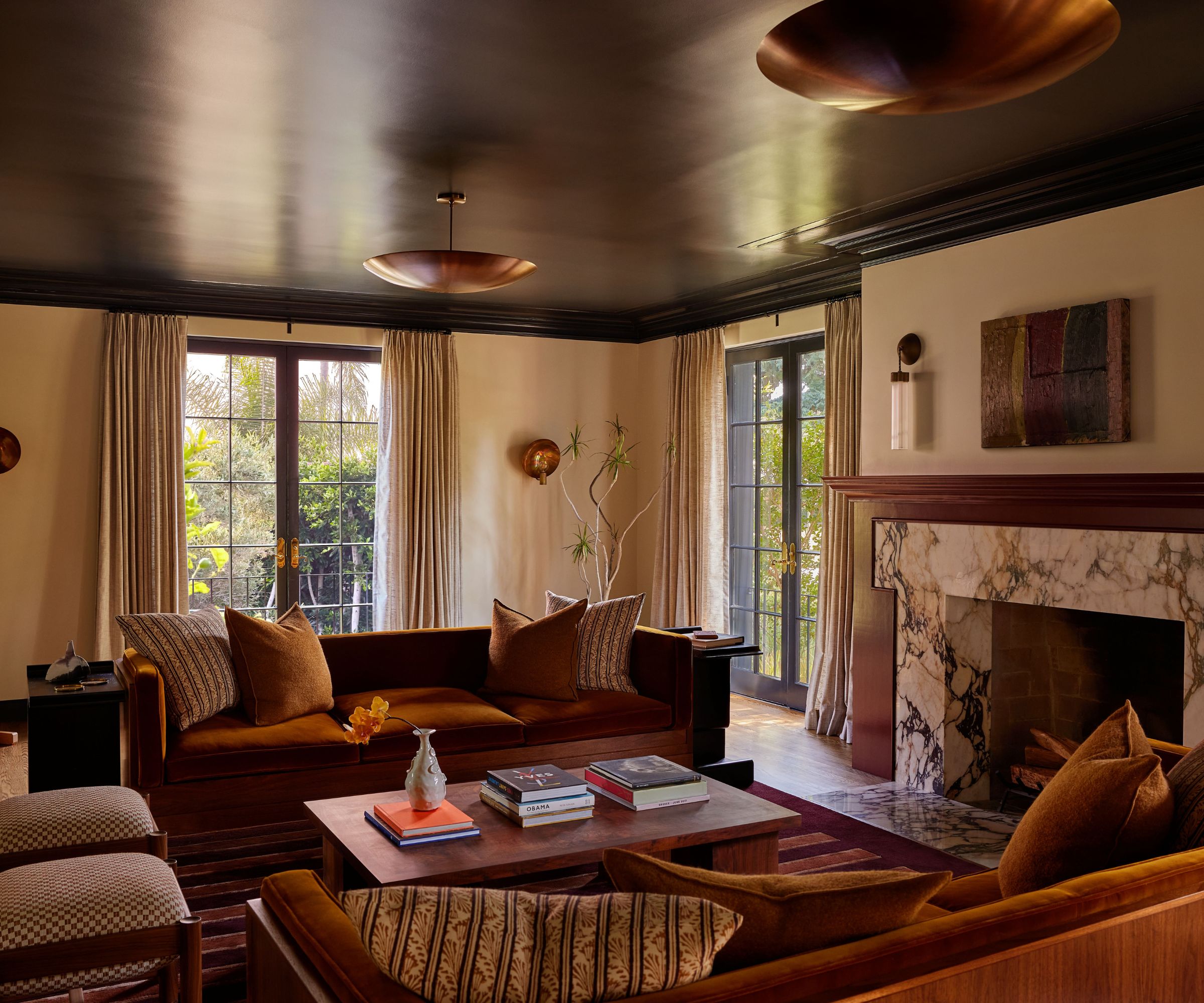 Living room with a brown-painted ceiling, cream walls, a large marble fireplace, two burnt-orange velvet sofas, a wooden coffee table, and an easel holding an oil painting in the corner.