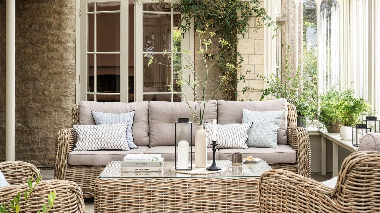 A sunroom with matching light wicker furniture set, including a three-seater sofa, two armchairs, and a glass-top table. 