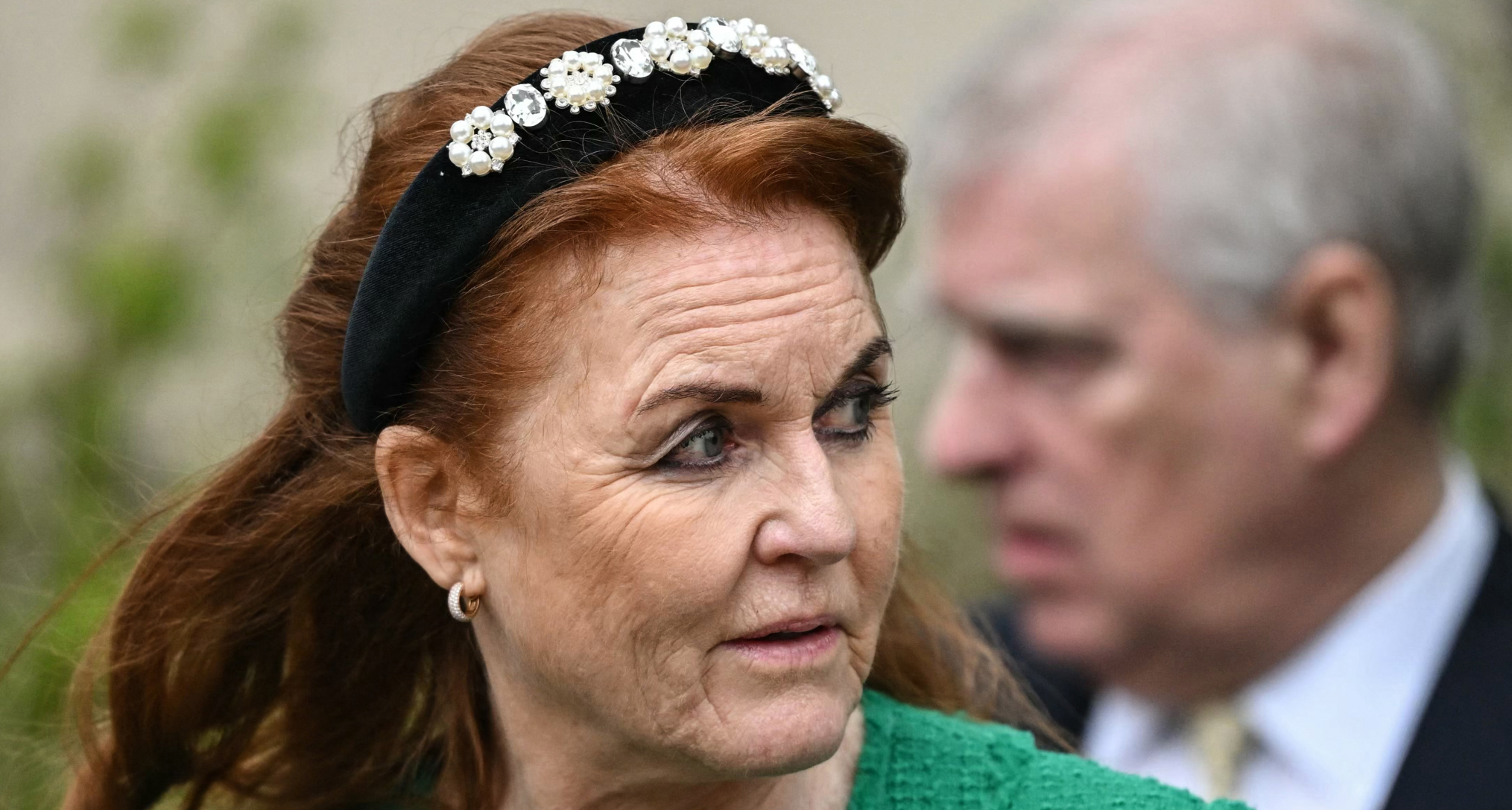 Sarah Ferguson wearing a jeweled headband and green coat looking to the side with Prince Andrew behind her