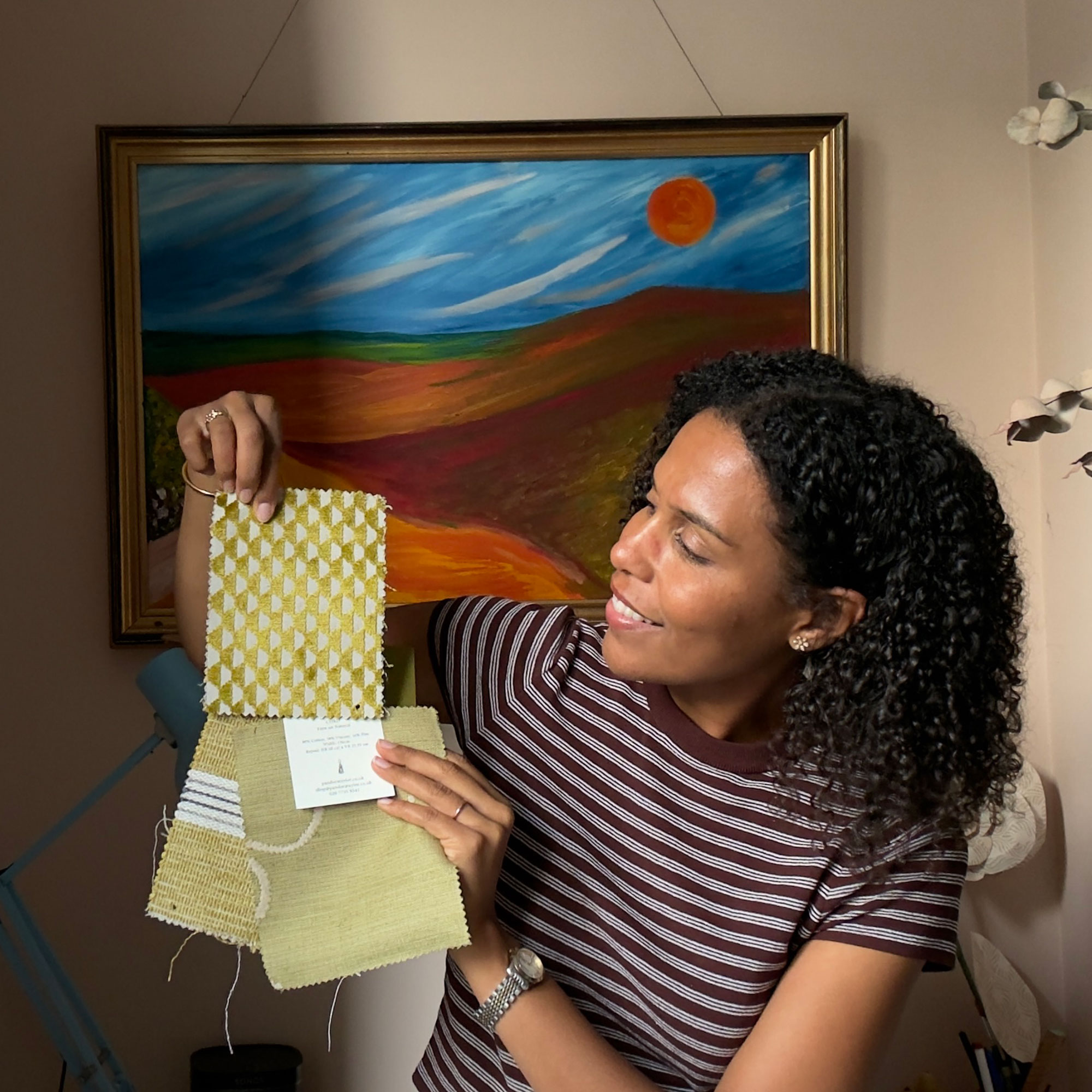 Woman in striped top holding fabric samples, sitting in front of landscape painting