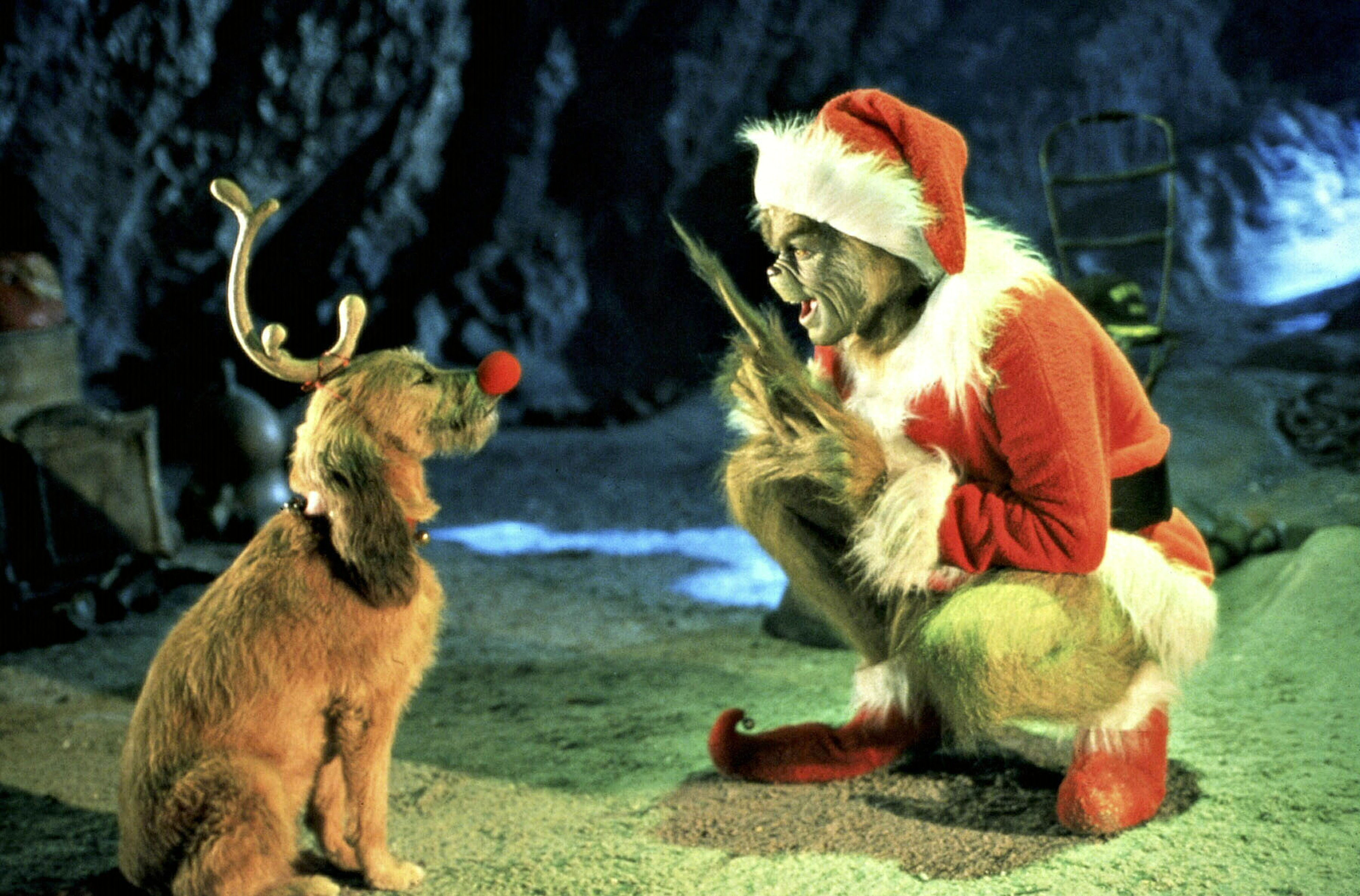 The Grinch, dressed in a Santa suit, crouches and talks animatedly to Max the dog, who sits wearing a single antler and a red Rudolph nose.