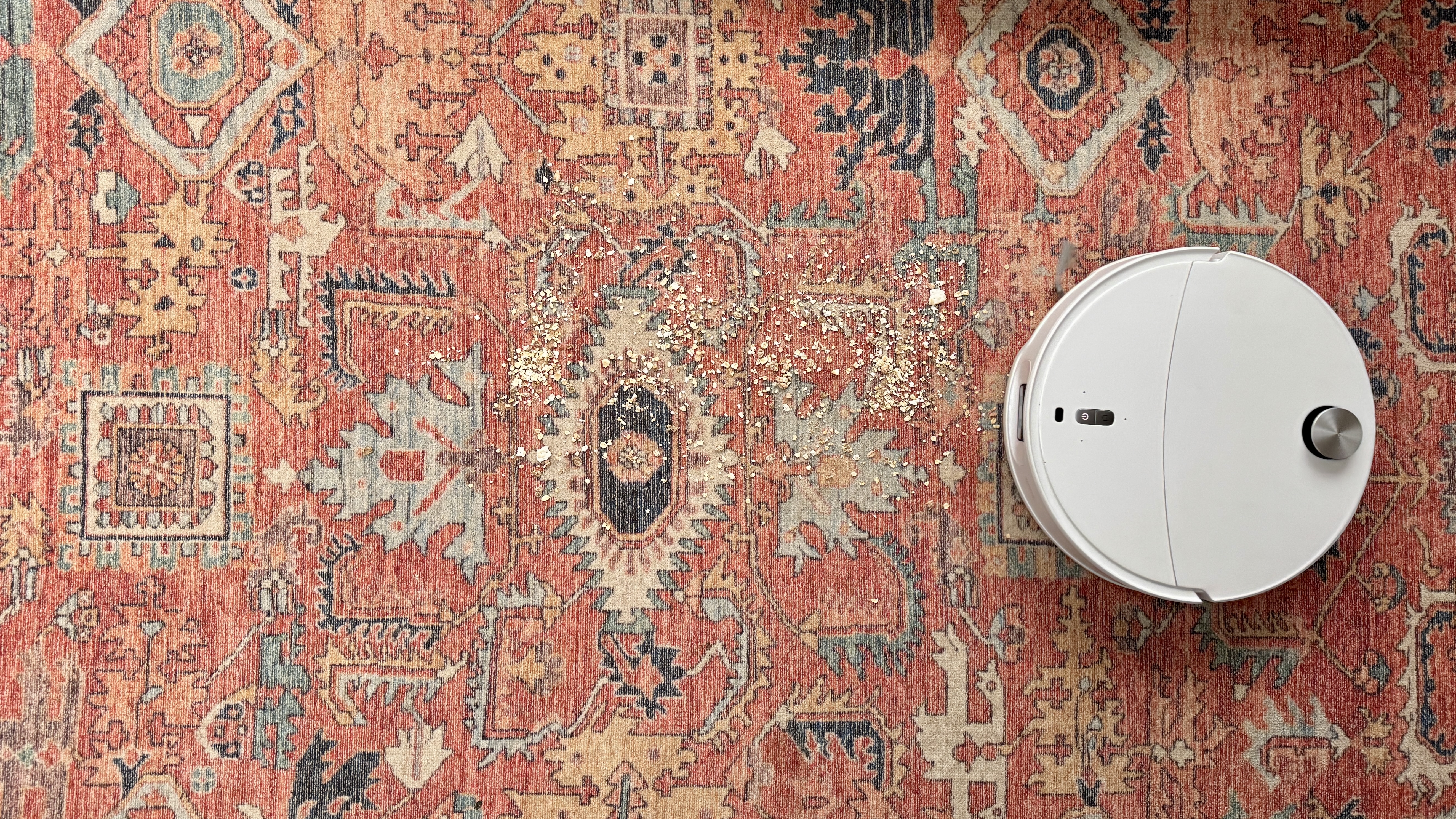 Roborock Qrevo Curv 2 Pro on a patterned carpet rug, with oats and crumbs sprinkled in front of it