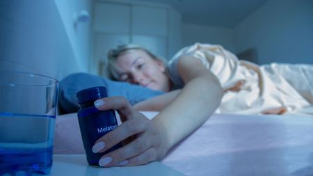 A woman in bed reaches for a blue bottle with the label melatonin on it on her nightstand