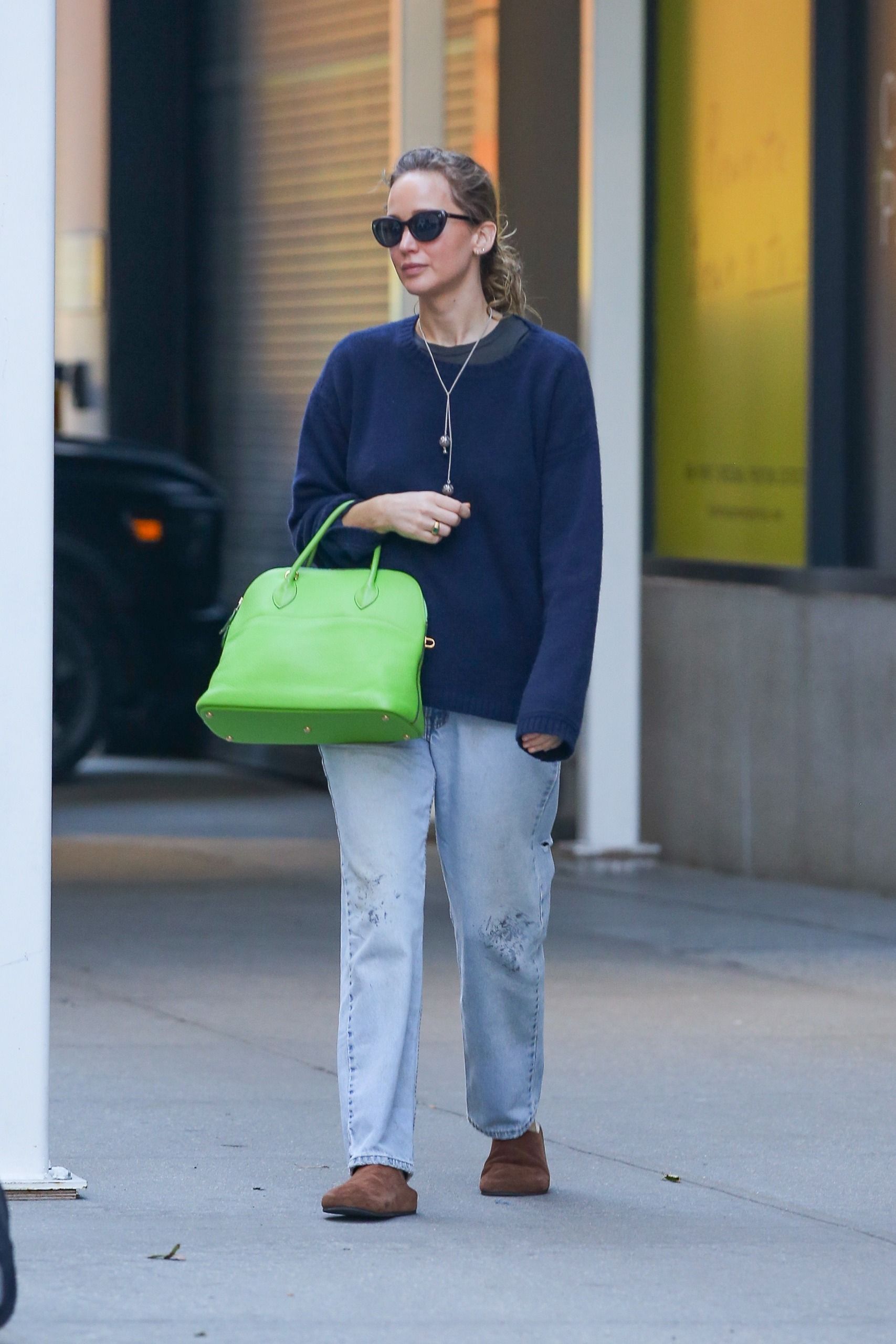 Jennifer Lawrence steps outside wearing a navy jumper with blue jeans, brown suede mules and an apple green leather bag.