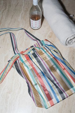 A stripy tote bag in pastel colors lays flat on a wooden surface next to a bottle of pink wine and a beige scarf rolled into a cylindrical shape.