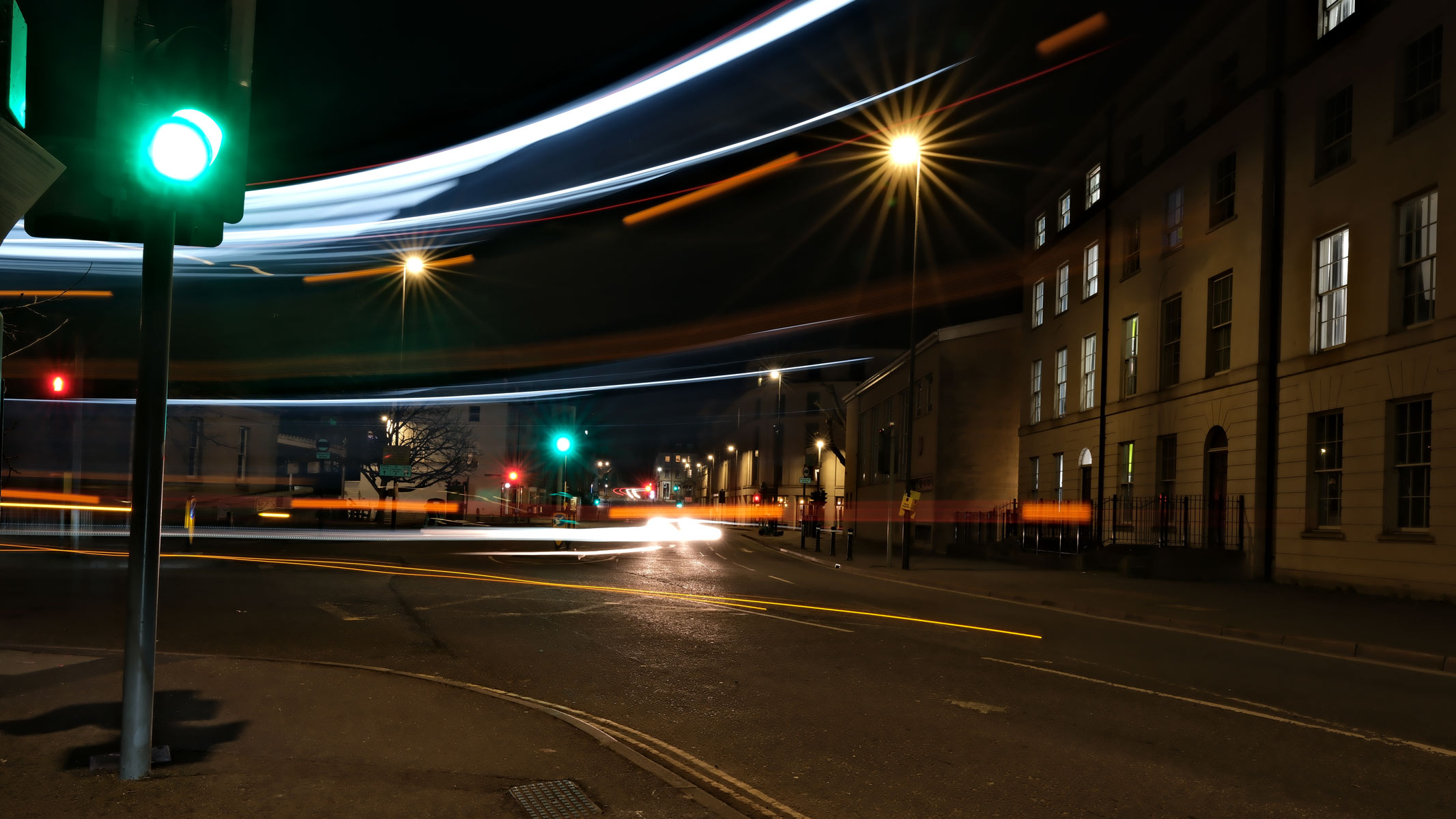 Light trails from a double-decker bus at night, on a street with apartment buildings, streetlights and traffic lights.