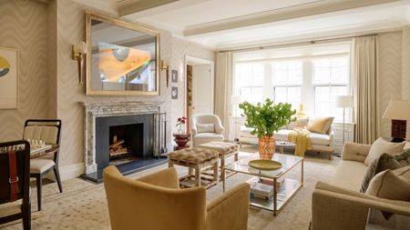 Bright living room with beige and cream interior, TV mounted on wall, sofas, armchairs, and a window bringing in daylight.
