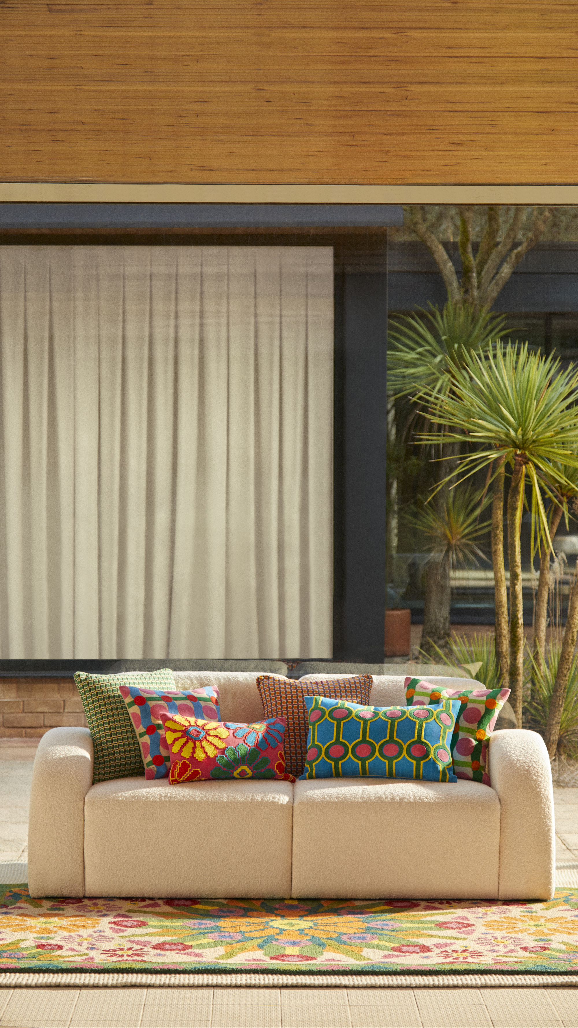 a curved white sofa with timber ceiling, white curtains, palms, colorful cushions on the sofa with botanic motifs, and a colorful rug