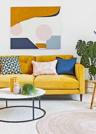 White living room with a mustard velvet sofa and tonal artwork hanging on the wall behind it to show how to decorate walls without painting in colours