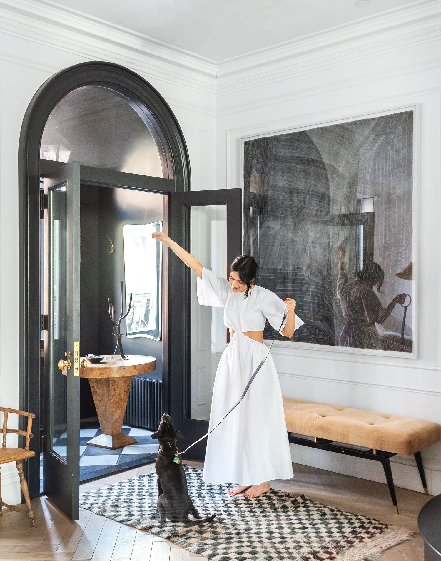 Athena Calderone stands in an entryway holding a lead attached to her sitting dog in front of a black-painted framed arched window and door and a large dark artwork on the white walls above the bench