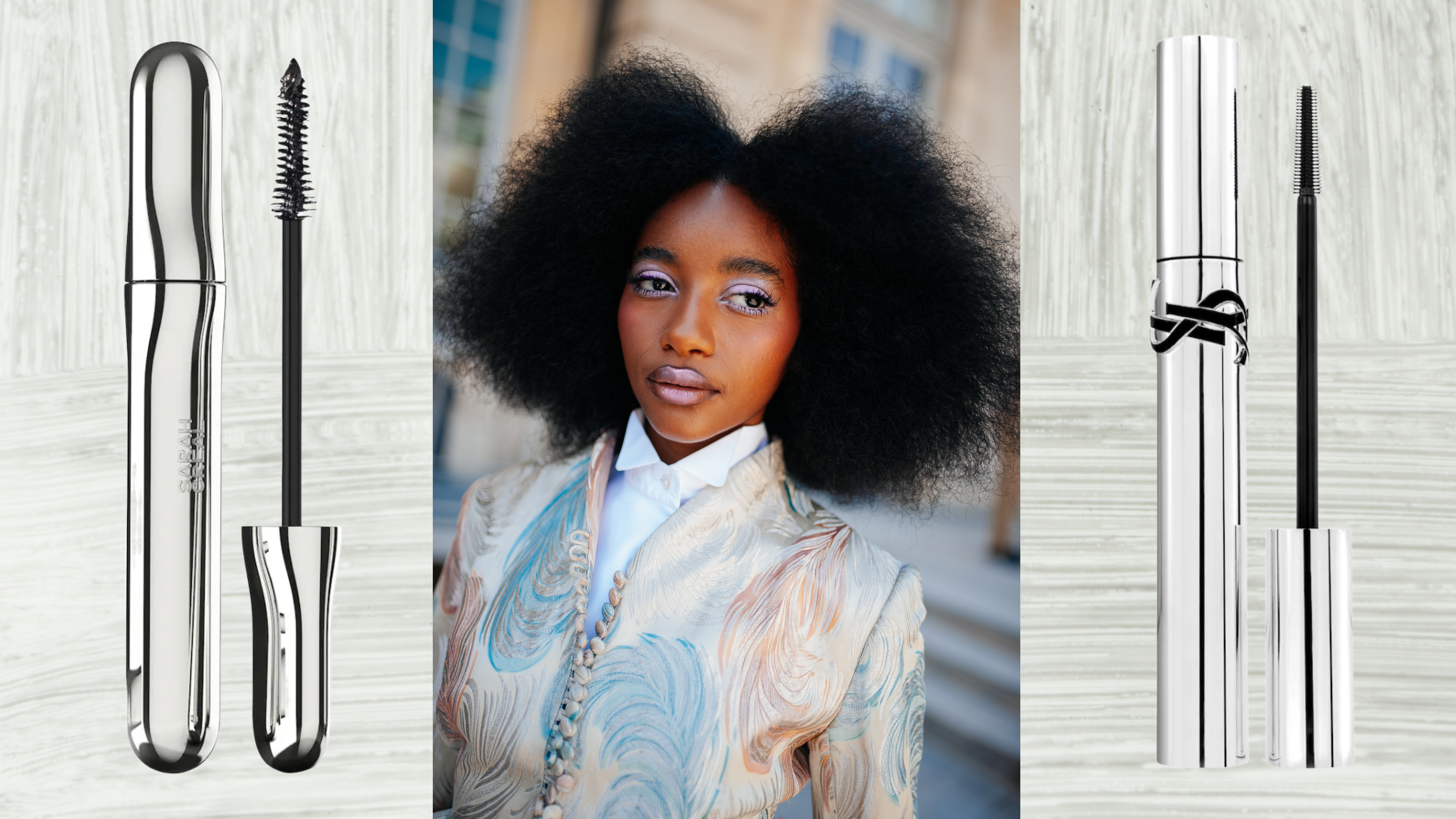 A woman wearing white eyeshadow with long lashes and her curls out in an afro. She is also wearing a pastel blazer and button-up shirt. She is next to two tubes of mascara. 