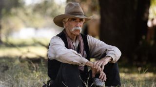 Sam Elliott sitting on the ground with his knees up and his arms resting on them in 1883. 