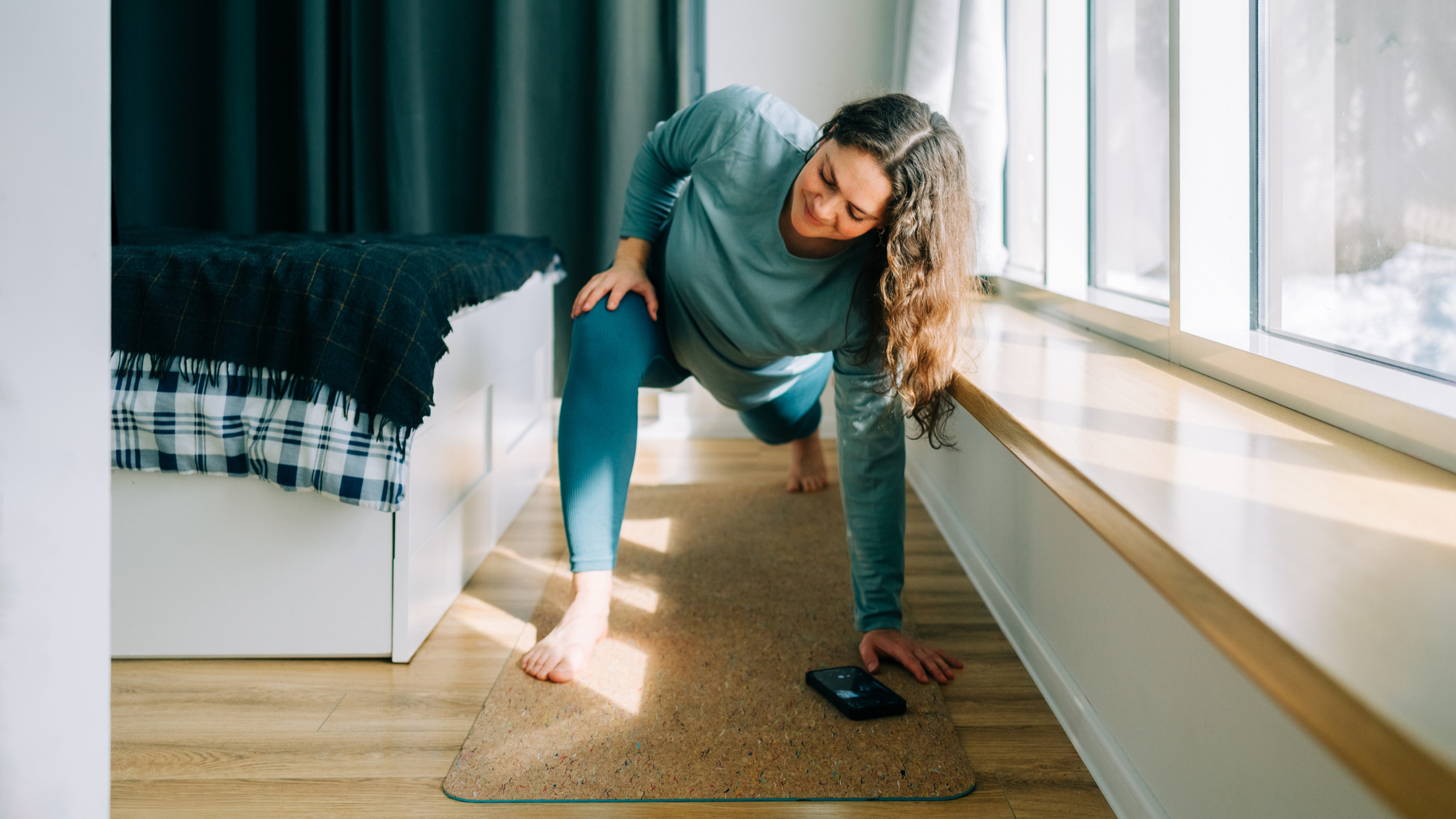 Woman stretching between a bed and a window, a smartphone is on the exercise mat in front of her