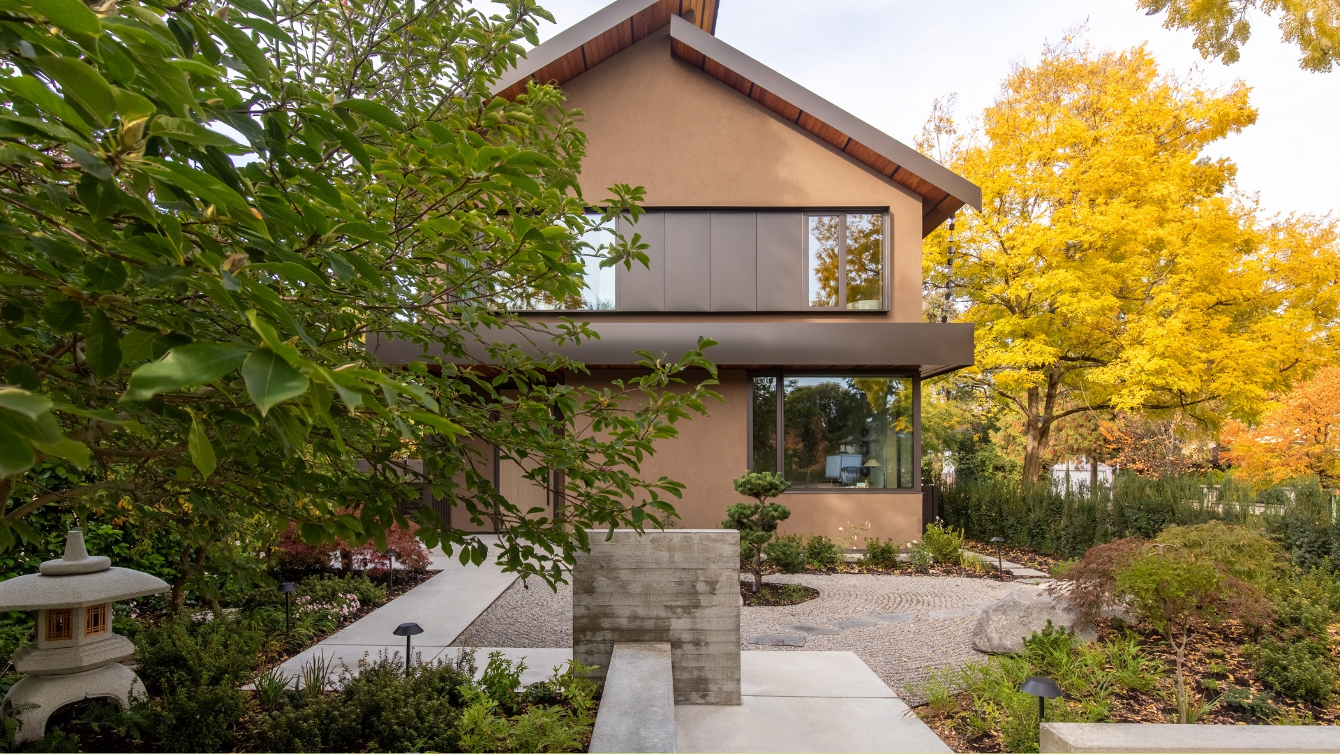 A Japanese garden with a bonsai, a gravel path, a stone lantern, and a two-storey home with a brown facade