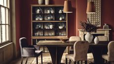 A deep red dining toom with a large wooden dining table. Upholstered chairs around it, and two hanging brown leather pendants above. A wooden cabinet of fine crockery in the background.