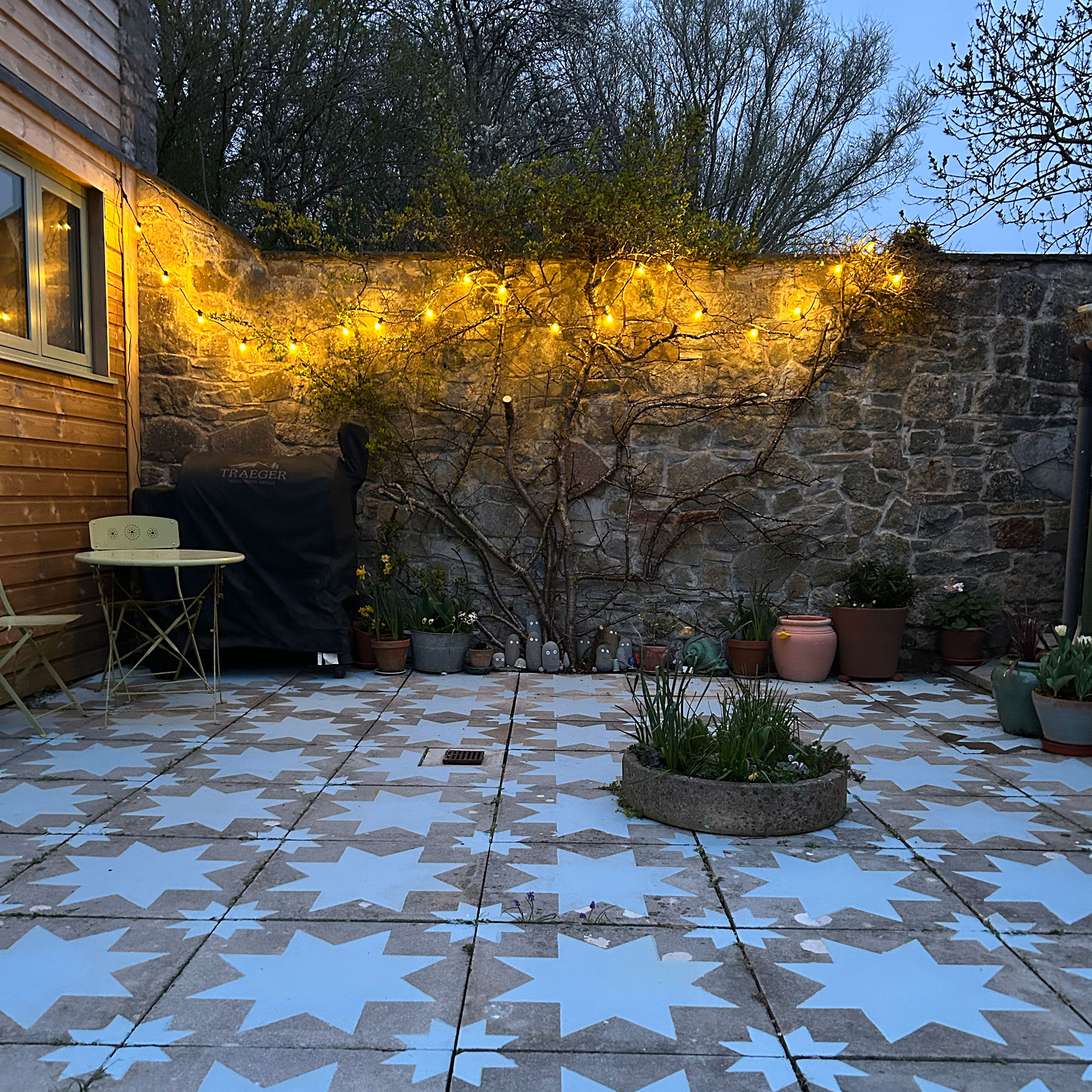 Stone wall enclosed patio in the evening with slabs painted with blue star design and with fairy lights on wall