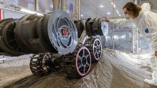 A box with multiple wheels rides over a bump in the dirt next to a man bending over.
