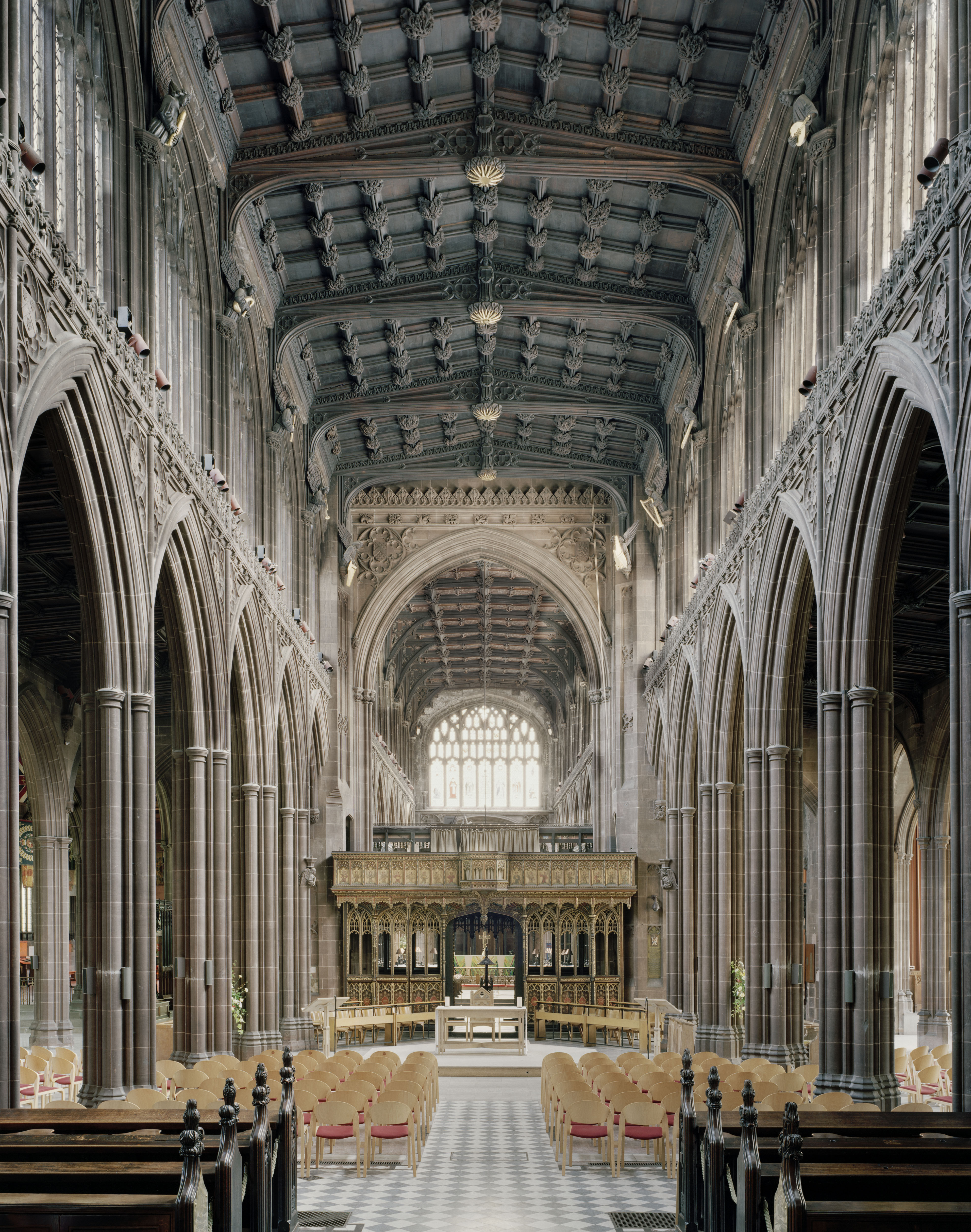 The nave of Manchester Cathedral, looking down rows of pale wooden chairs towards a gilded rood screen beneath a dark carved hammerbeam roof.