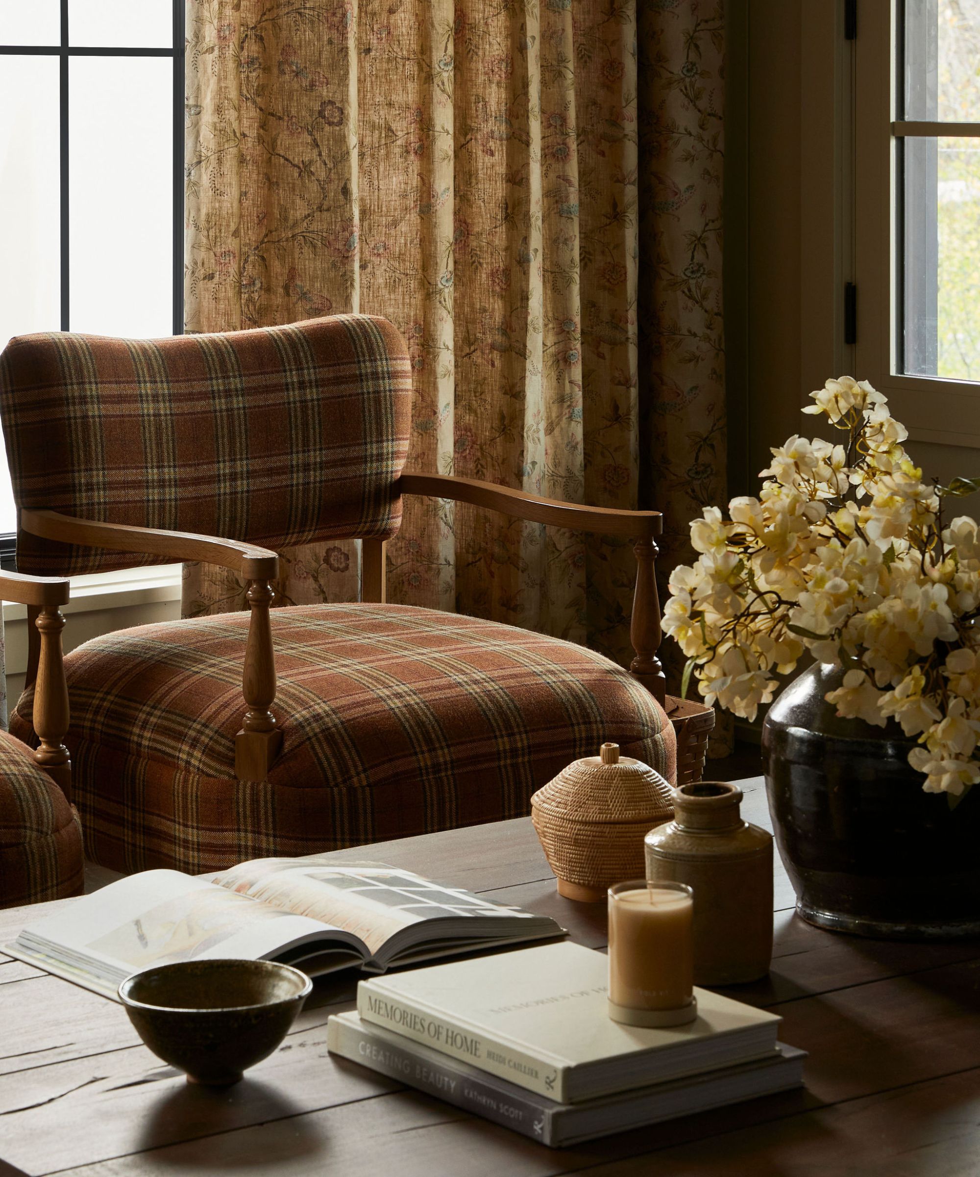 Close up of check armchair and wooden coffee table decorated with books, candles, and a vase of flowers, floral patterned curtains in background