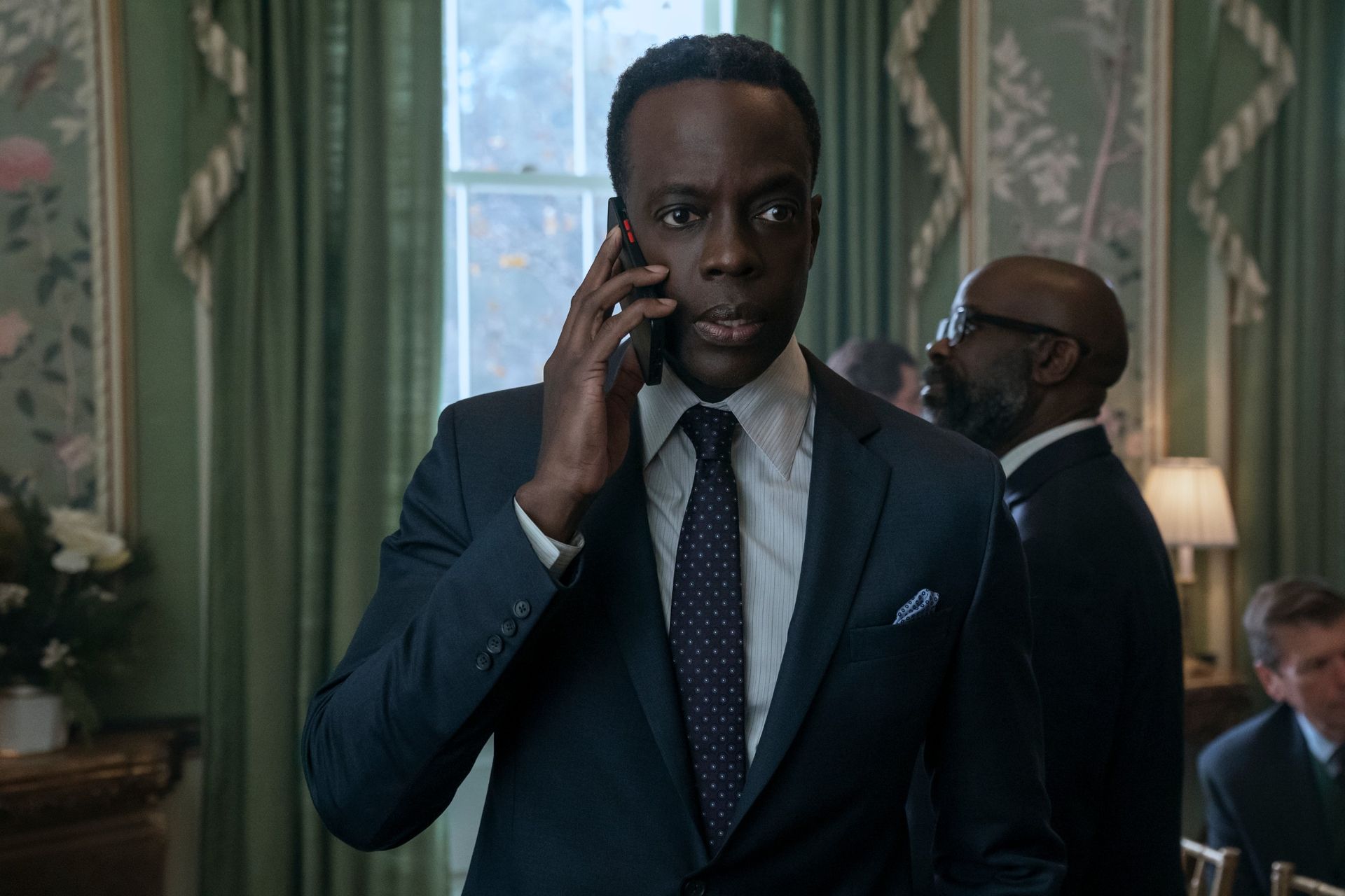 Ato Essandoh as Stuart Hayford, holding a phone to his ear while standing in a crowded room in front of windows with green curtains, in episode 402 of The Diplomat.
