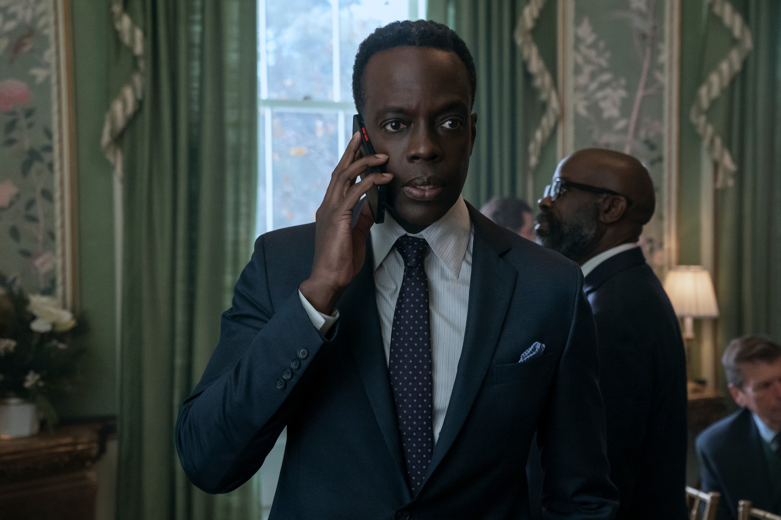 Ato Essandoh as Stuart Hayford, holding a phone to his ear while standing in a crowded room in front of windows with green curtains, in episode 402 of The Diplomat.