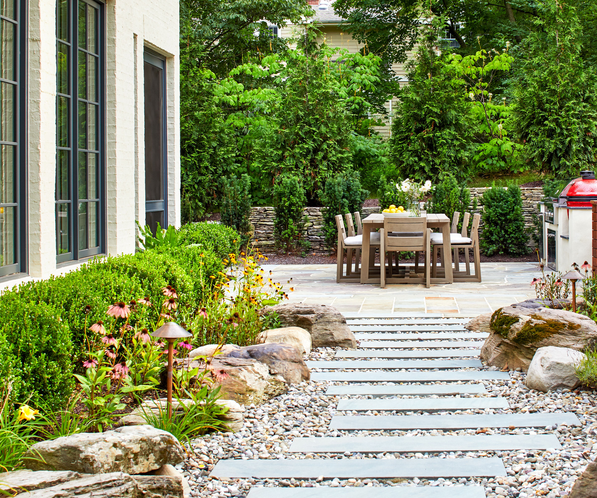path made of pavers and gravel, with rocks, plants and low hedge, and garden furniture set with evergreen trees in the background