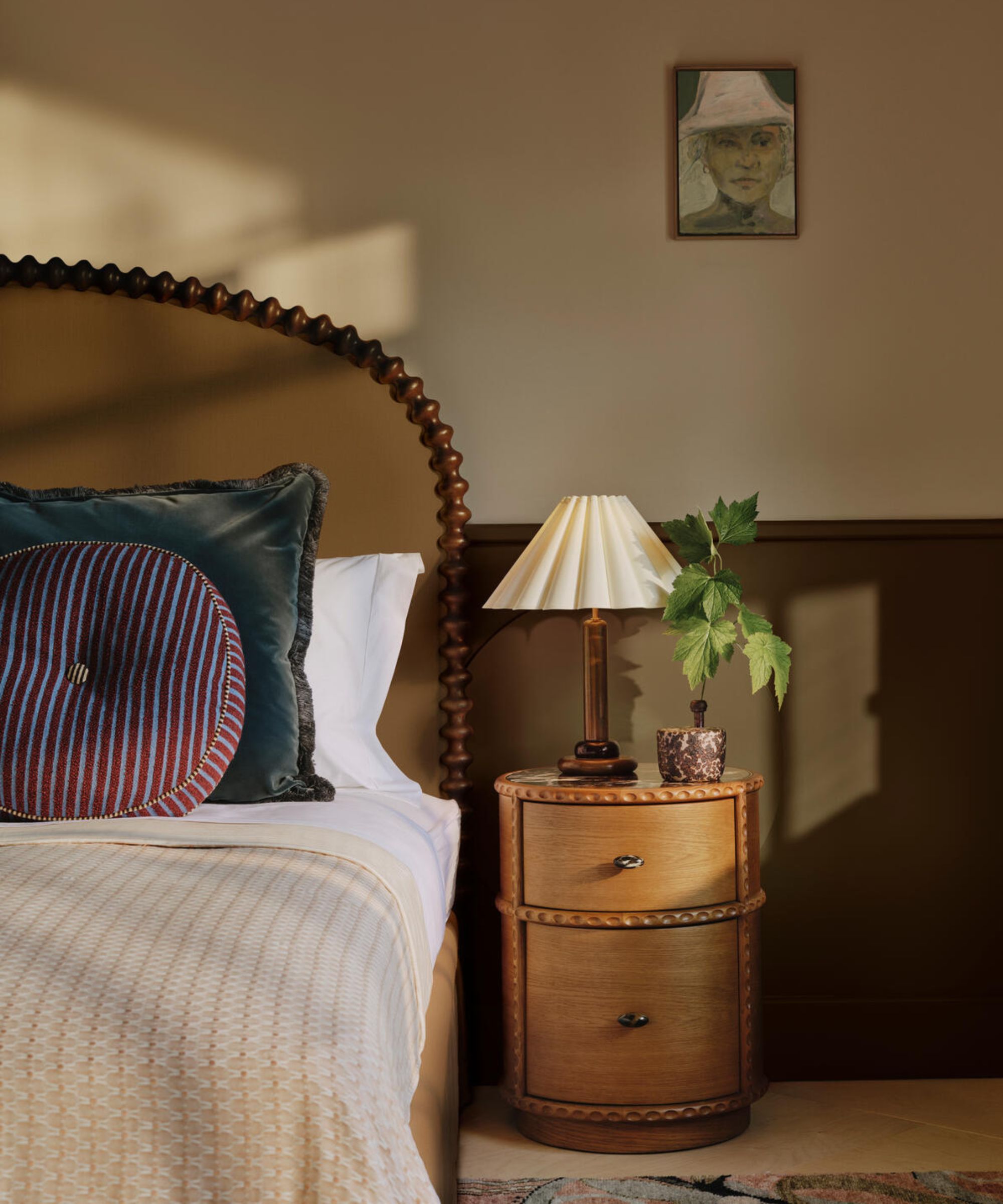Bedroom styled by Soho Home including a round bedside table with lamp and potted plant, curved bobbin bedhead with bed dressed with white linen, beige throw and blue cushions, and an artwork on the wall
