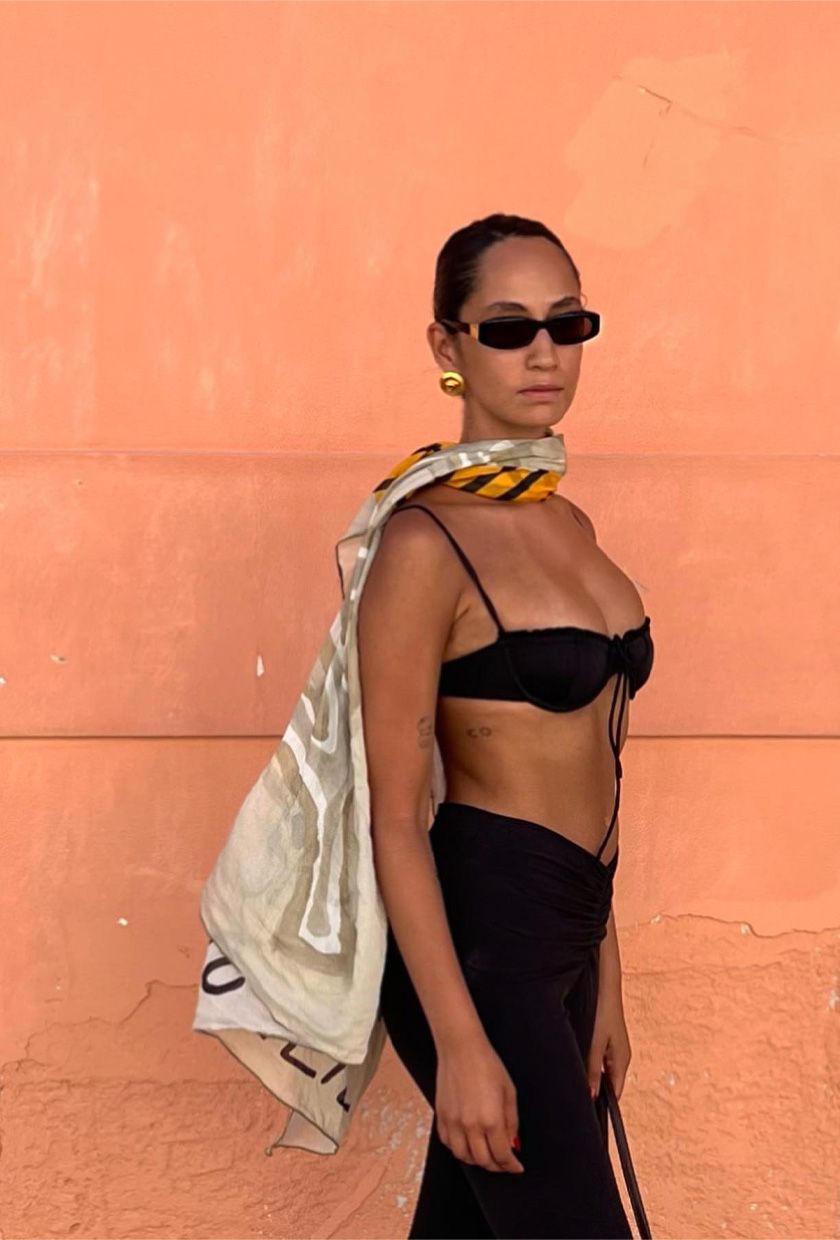 classic swim trend shown on a French woman standing outside wearing a black balconette bikini top with black capri pants, a printed scarf around the shoulders, gold earrings, and black sunglasses