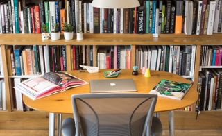 Grey chair and curved-edge wooden desk with books, an Apple MacBook laptop and a stapler. Behind the desk is a filled bookcase
