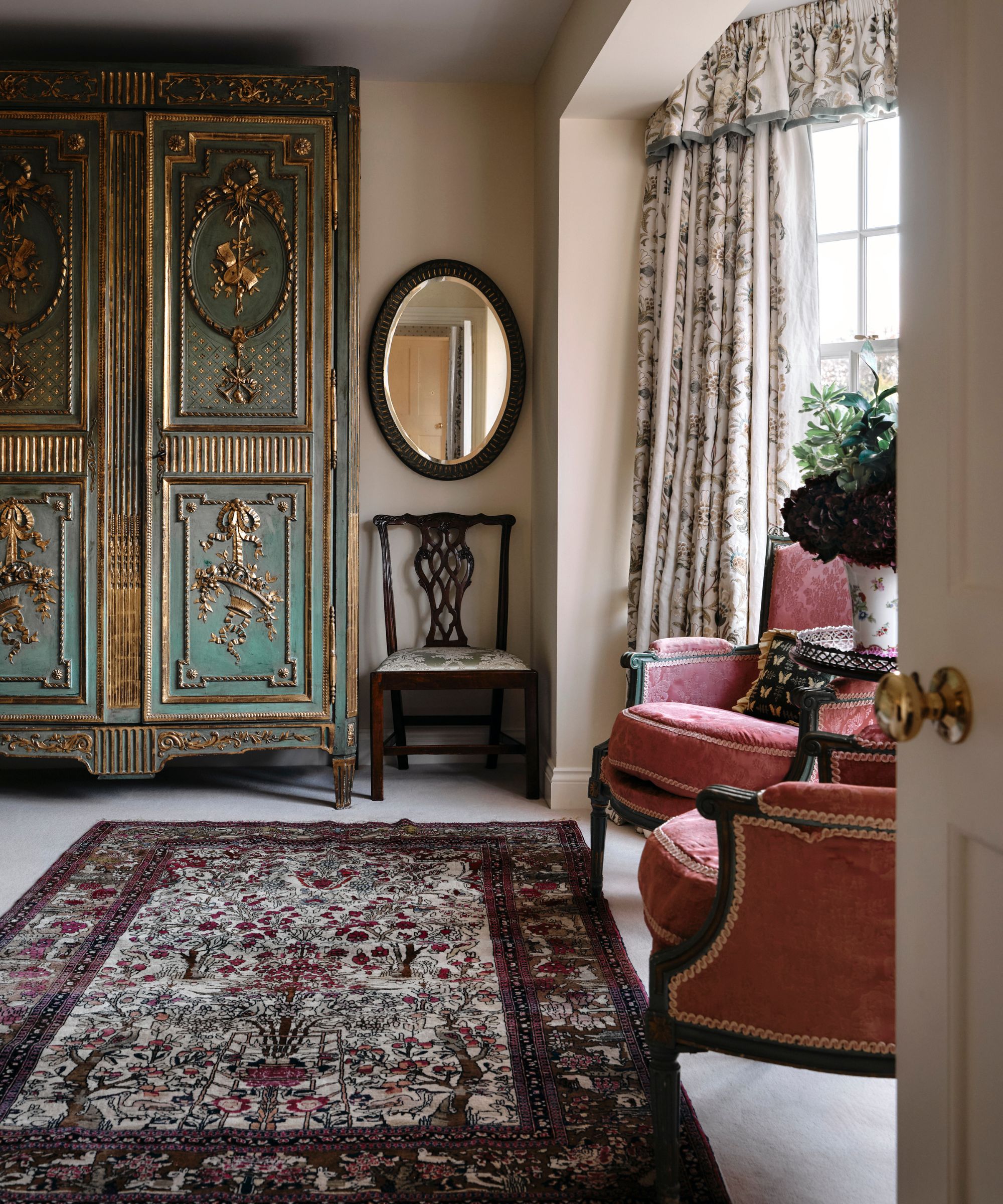 Traditional living room with pale pink walls, persian rug on floor, antique blue armoire, two pink antique armchairs, dark wooden mirror and large window with pelmet curtains
