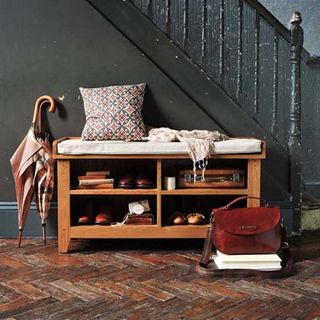 Oakland Shoe Storage Bench and Cushion containing a selection of shoes, books and bags, with umbrellas next to it and in front of a grey staircase
