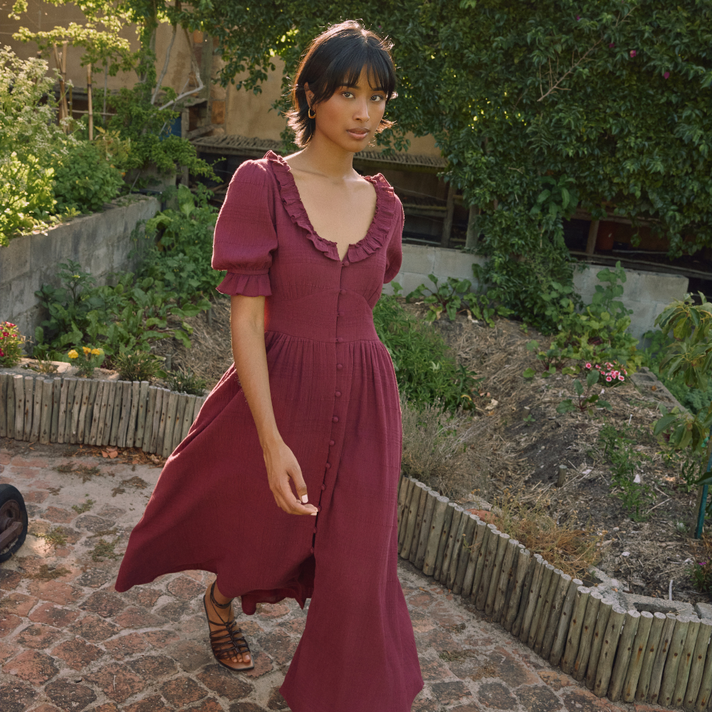 Model walking through a paved garden wearing a plum maxi dress and sandals sold at Nobody's Child