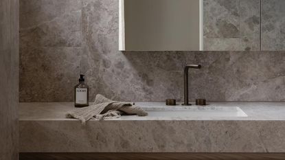 A bathroom with a stone countertop and backsplash as well as a sink with dark brass hardware and a mirror above the sink