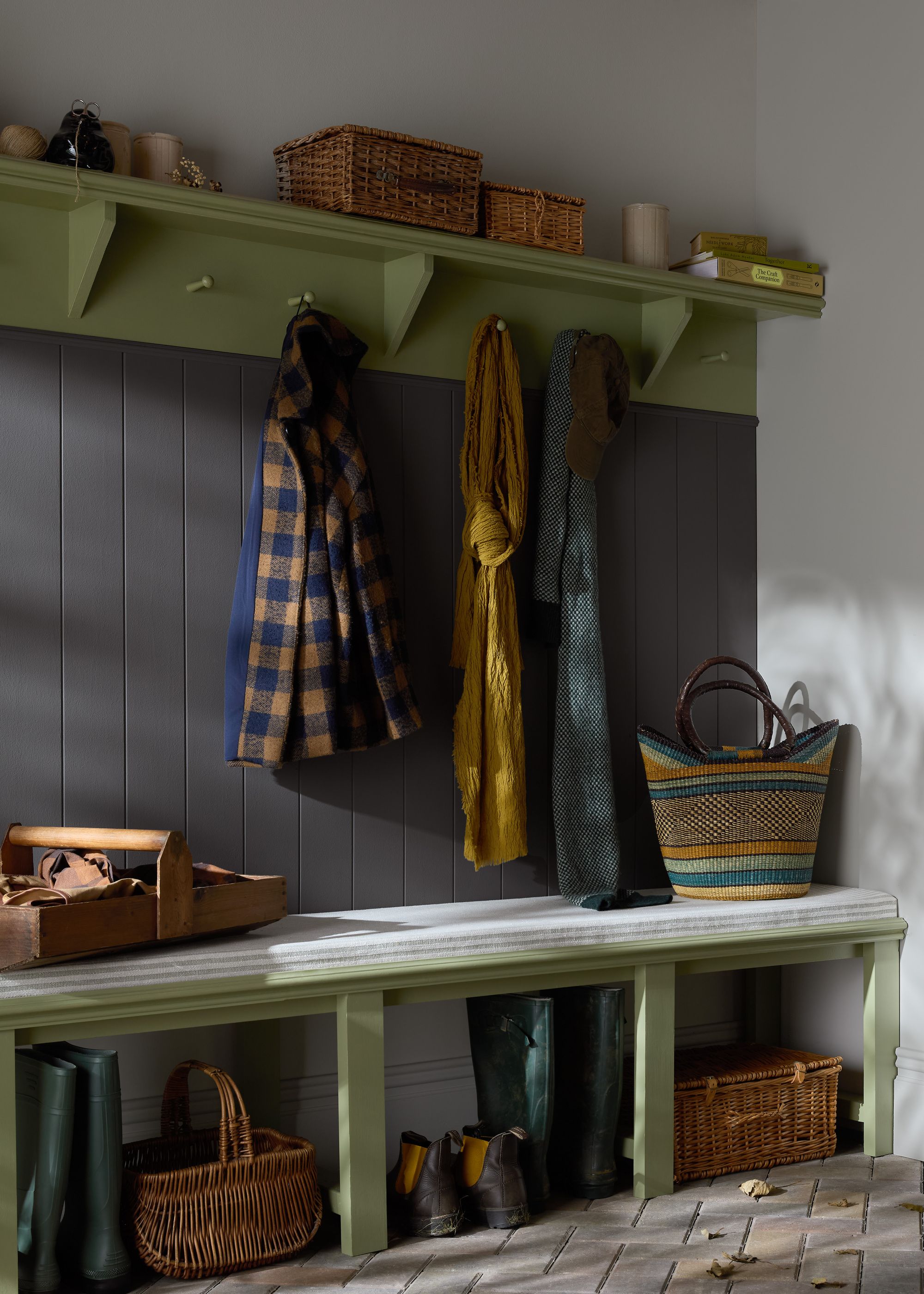 Angled image of a mudroom with white walls, dark gray paneling, and light sage green shelving and trim. Coats and scarves are hanging from the hooks on the built-in, and baskets and shoes are under the built-in.