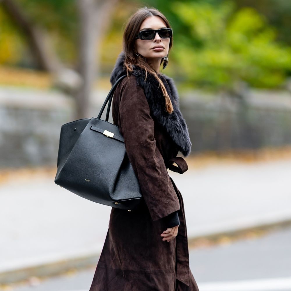 Emily Ratajkowski wears a brown suede trench coat with a fur collar. She styles her coat with black trousers and black kitten heel boots.