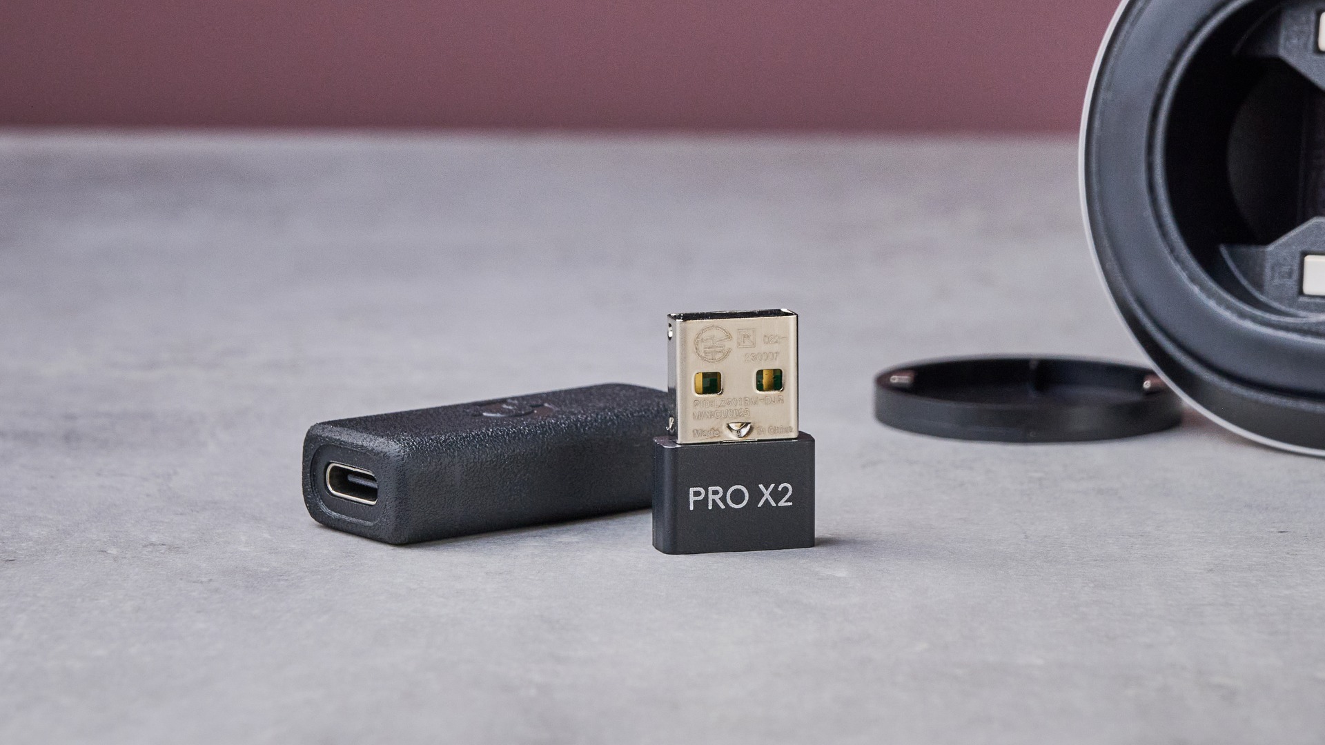 USB dongle and extender for Logitech G Pro X2 Superstrike, with Logitech G Pro X2 Superstrike in background with underside lid removed, on white desk with pink wall in background