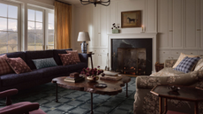 Moody living room featuring a blue geometric rug, navy sofa, floral love seat, live wood coffee table, beige drapery, vintage-inspired throw pillows, and equestrian art overhead the fireplace