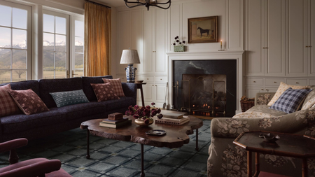 Moody living room featuring a blue geometric rug, navy sofa, floral love seat, live wood coffee table, beige drapery, vintage-inspired throw pillows, and equestrian art overhead the fireplace