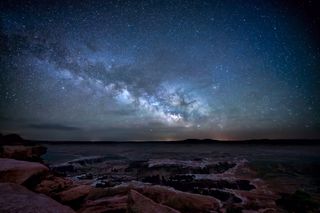 the milky way stretches across a star-studded sky with rocky terrain below.