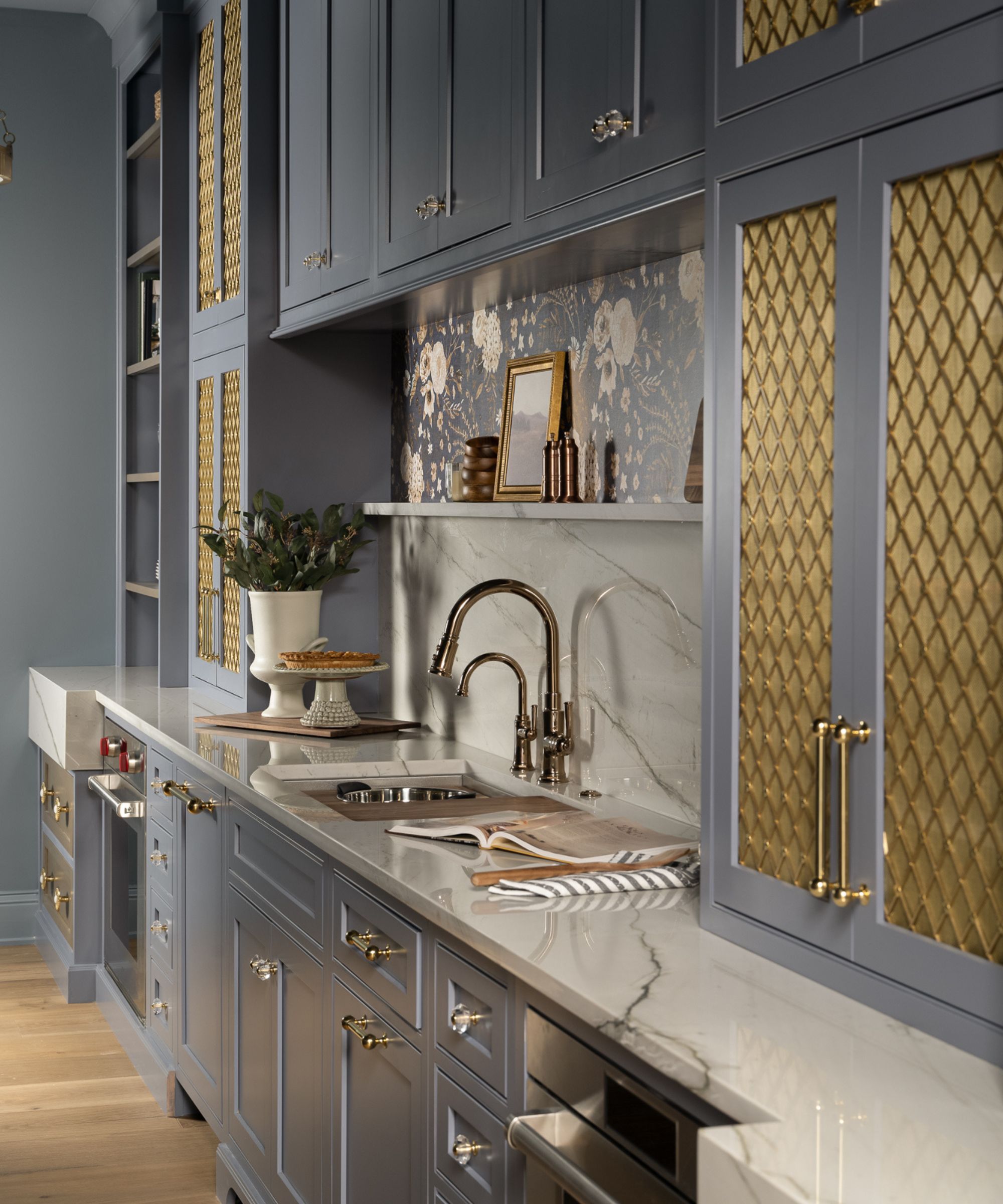 A close-up of a blue-gray butler's pantry featuring gold mesh cabinet inserts, a marble countertop with an integrated sink, and a floral patterned backsplash.