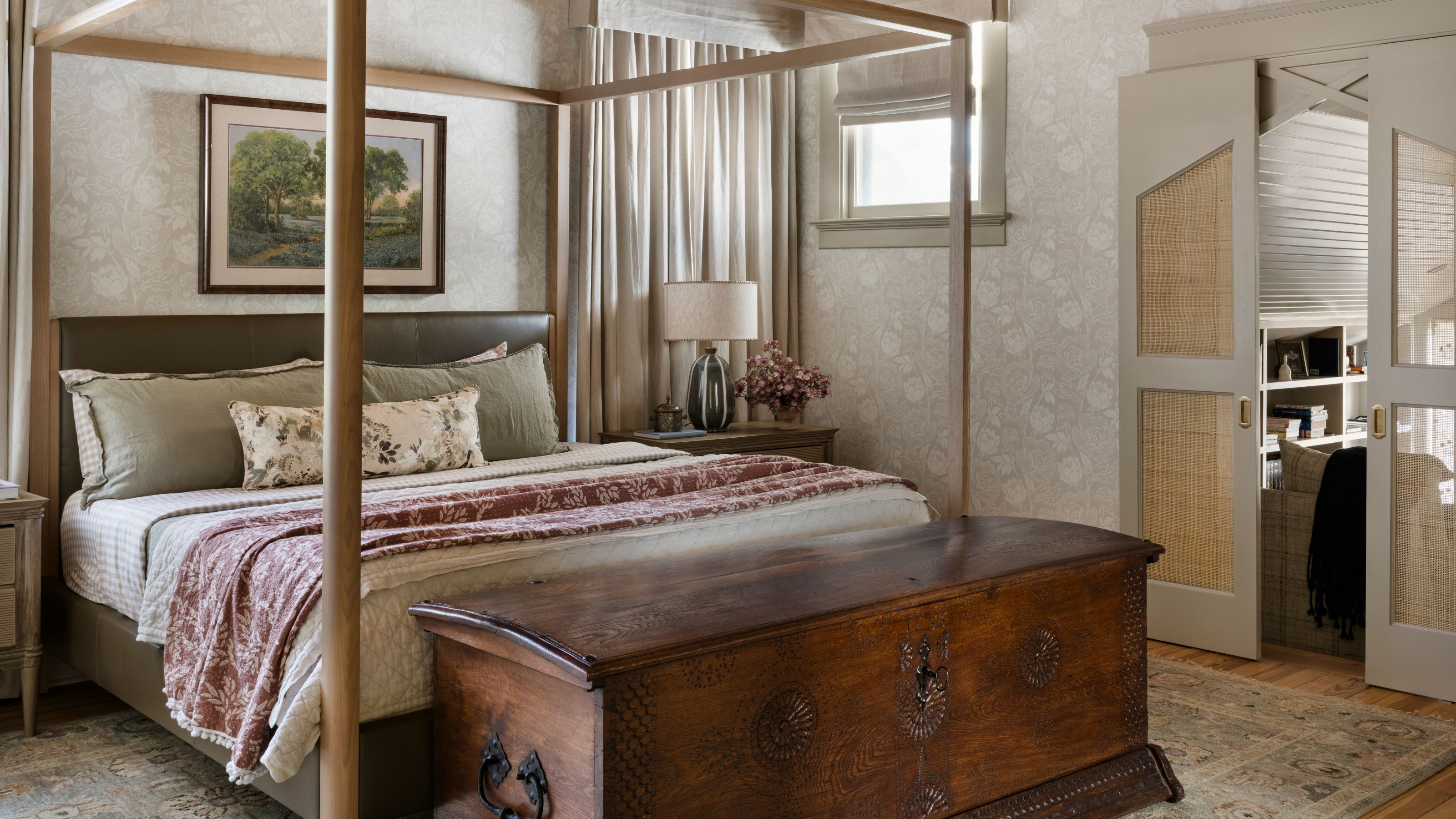 a muted green primary bedroom with floral wallpaper, a four poster bed, layered linens and a large dark wooden antique chest at the foot of the bed
