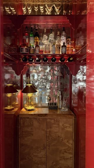 Image of a small closet that has been renovated into a home bar. The walls are a glossy, bright red, and there is a wooden cabinet and a high shelf full of bottles and glasses